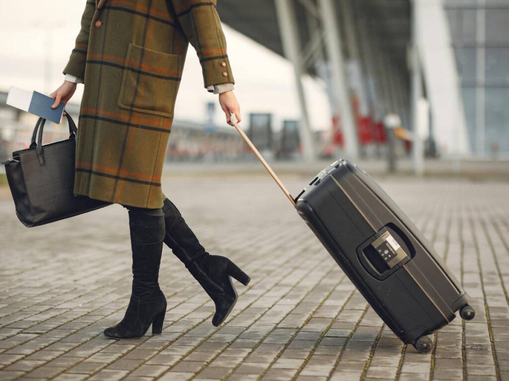 Stylish woman with suitcase and bag walking on street near modern airport terminal