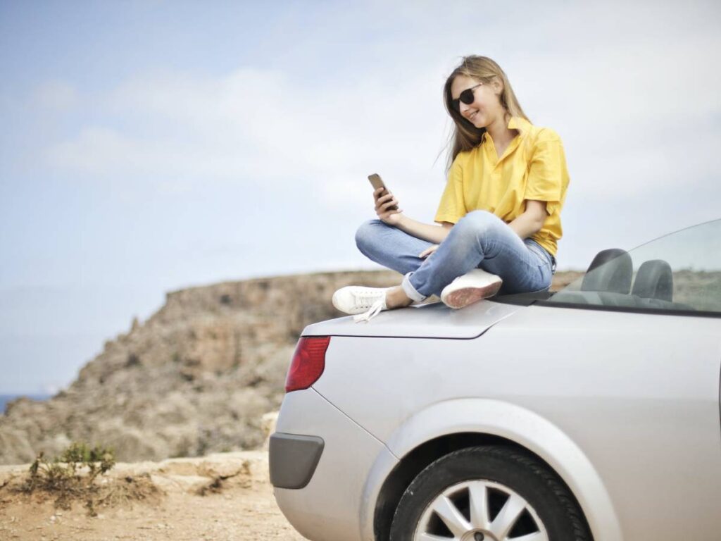 Woman in Yellow Blouse and Blue Jeans Taking Selfie While Sitting on Car